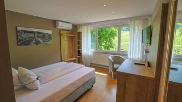 A bedroom with a bed, a desk, a chair, a TV, and a window with a view of greenery.