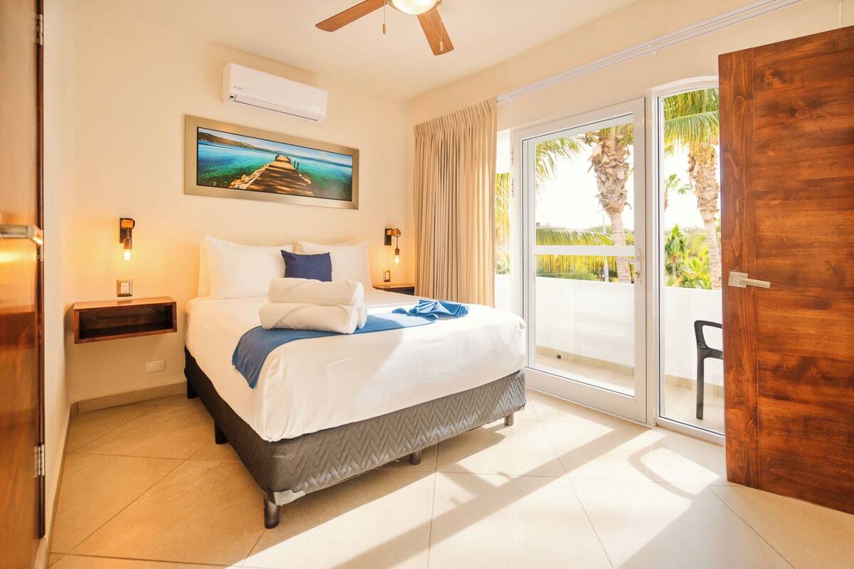 Una habitación de hotel con cama, mesita de noche, unidad de aire acondicionado, ventilador de techo, puerta corrediza al exterior y un cuadro en la pared.