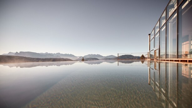 Un edificio moderno con fachada de vidrio que refleja un lago tranquilo y montañas distantes.