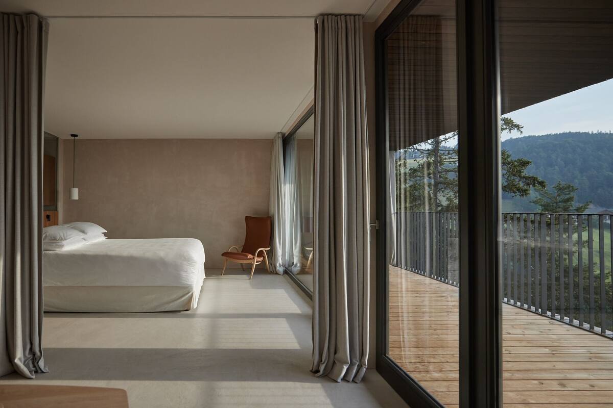 Une chambre moderne avec un grand lit, une chaise et une vue sur un balcon doté d’une terrasse en bois et des montagnes en arrière-plan.