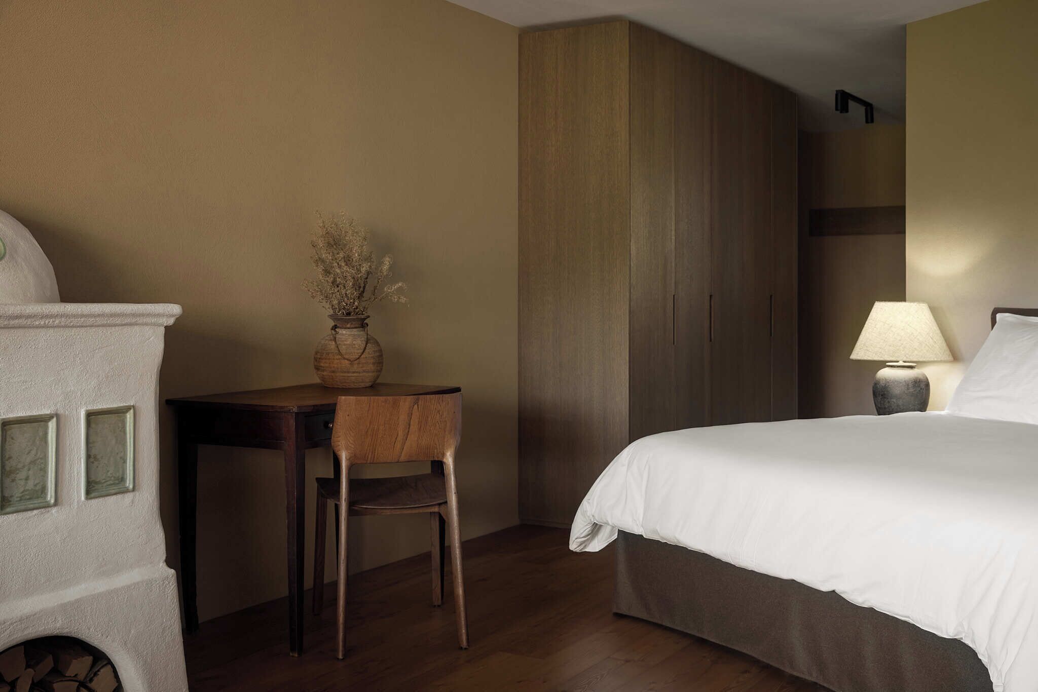 A hotel room with a bed, a desk with a chair, a vase with dried flowers, and a lamp.