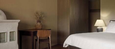 A hotel room with a bed, a desk with a chair, a vase with dried flowers, and a lamp.