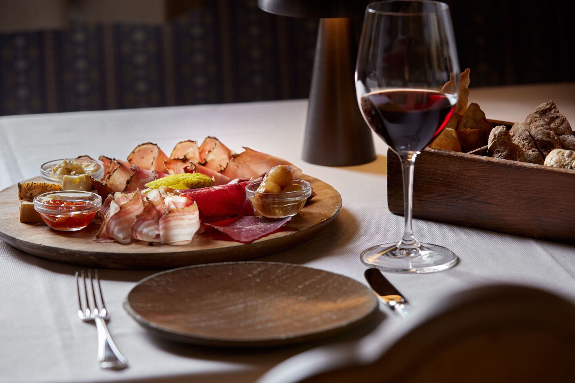 Une planche en bois garnie de différentes sortes de charcuteries et d’accompagnements, un verre de vin rouge et un décor rustique.