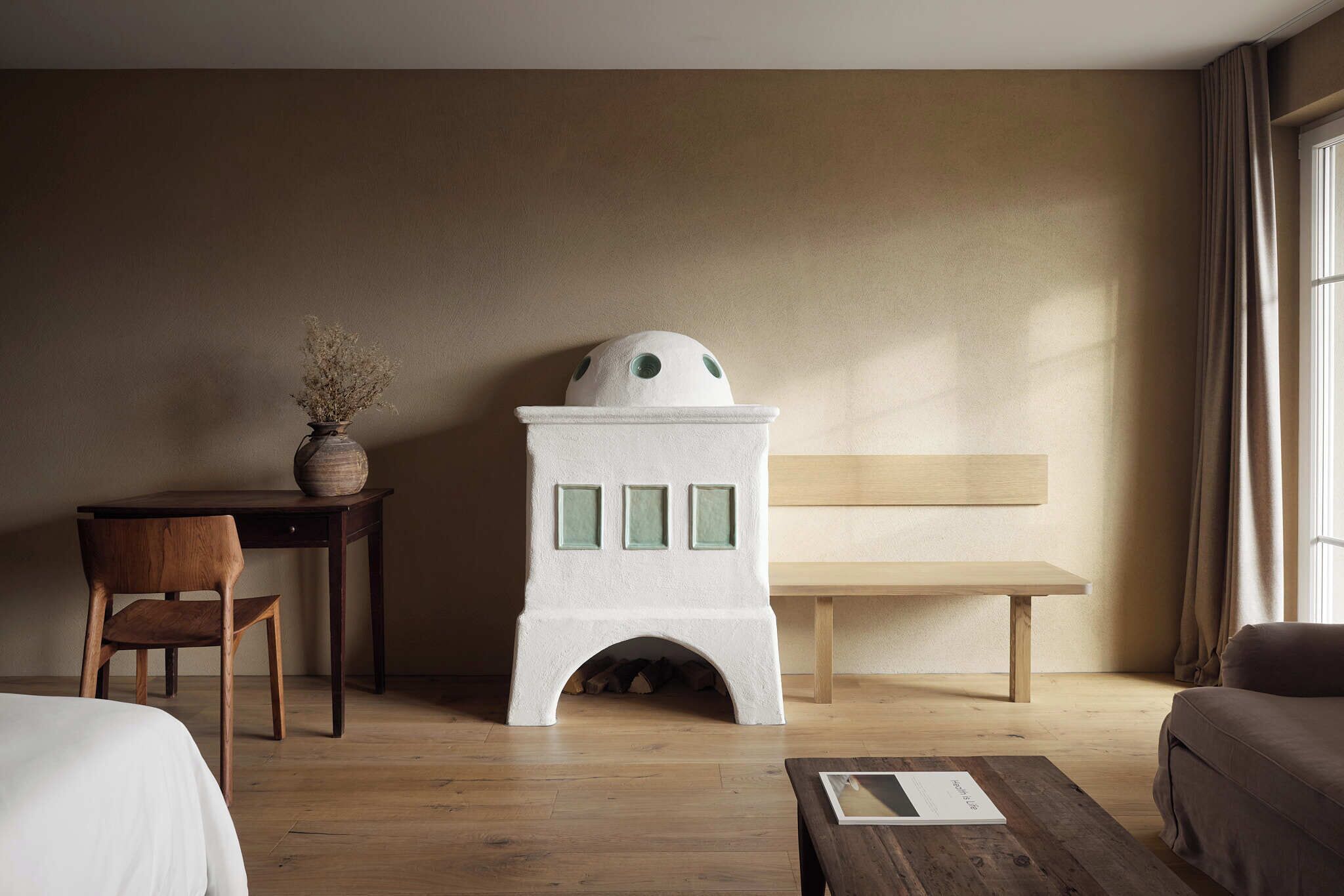 Una habitación con escritorio de madera, una silla, chimenea blanca, un banco y una cama.