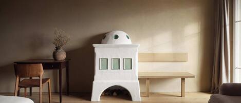 A room with a wooden desk, a chair, a white fireplace, a bench, and a bed.