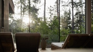 Una habitación con un ventanal grande con vistas a un bosque, dos sillas de madera y una mesita con un jarrón.