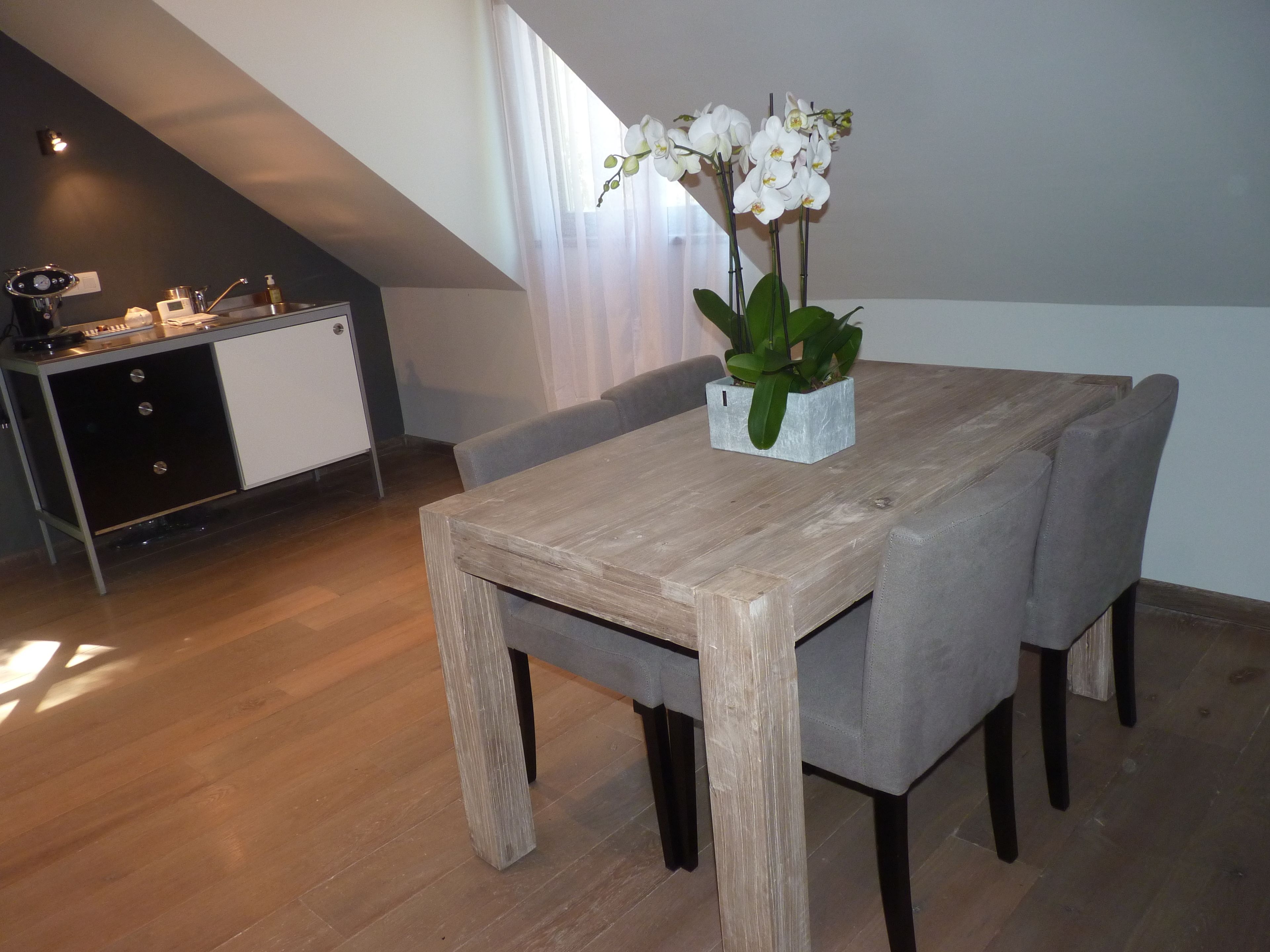 A dining area with a wooden table and grey upholstered chairs, a potted orchid on the table, and a kitchen in the background.