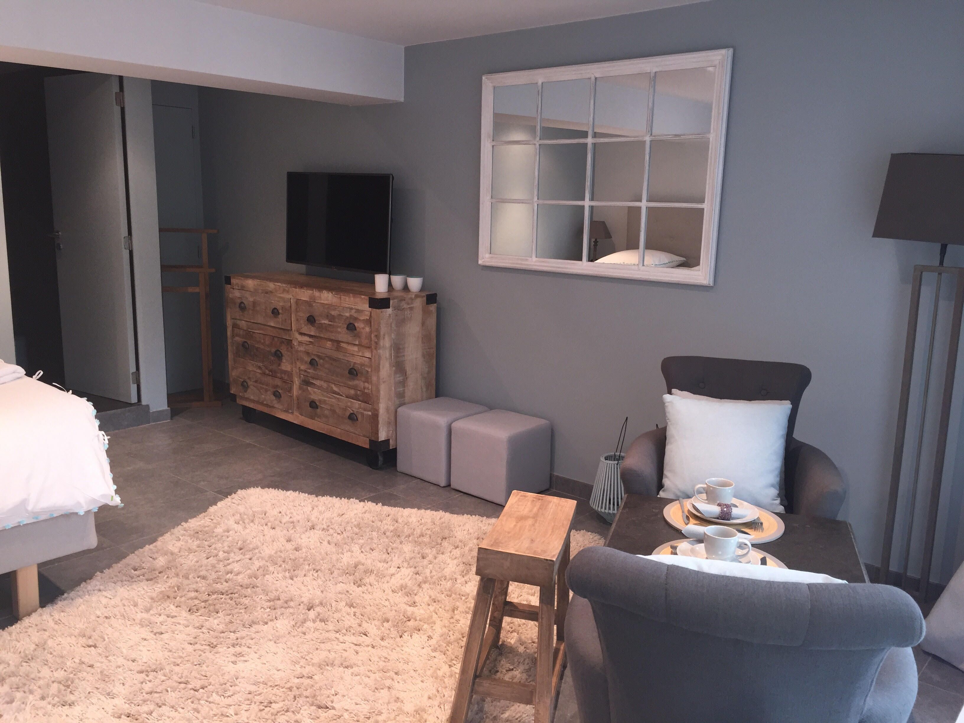 Een moderne slaapkamer met een bed, een houten dressoir, een televisie, een raam en een zithoek met een tafel en stoelen.
