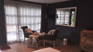 Una habitación con una cama, dos sillas, una mesa con tazas, una lámpara, un ventanal con cortinas y un aparador.