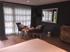 Una habitación con una cama, dos sillas, una mesa con tazas, una lámpara, un ventanal con cortinas y un aparador.