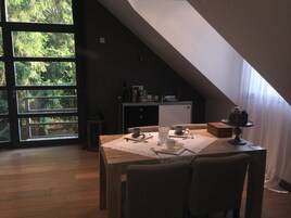 Un comedor con una mesa preparada para dos personas, una cocina al fondo y vistas a árboles a través de amplios ventanales.