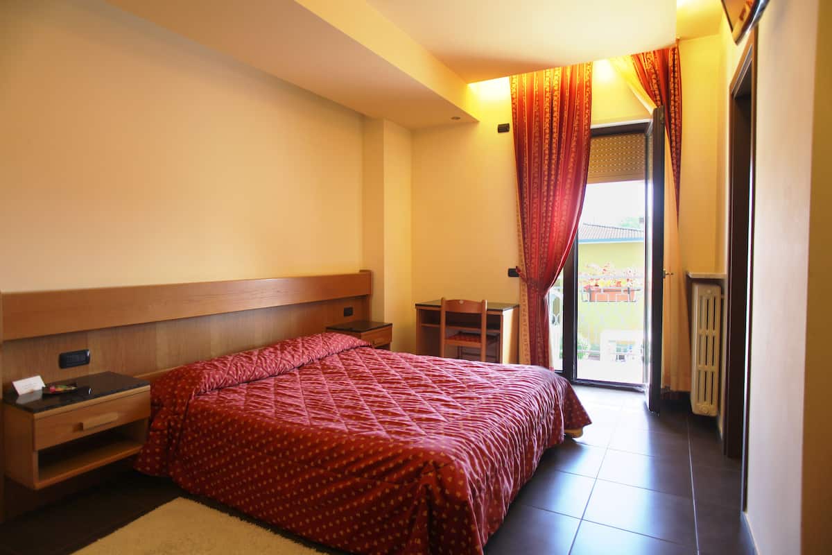 Una camera da letto con un letto, una scrivania e una sedia. Si vede una finestra con tende e un termosifone.