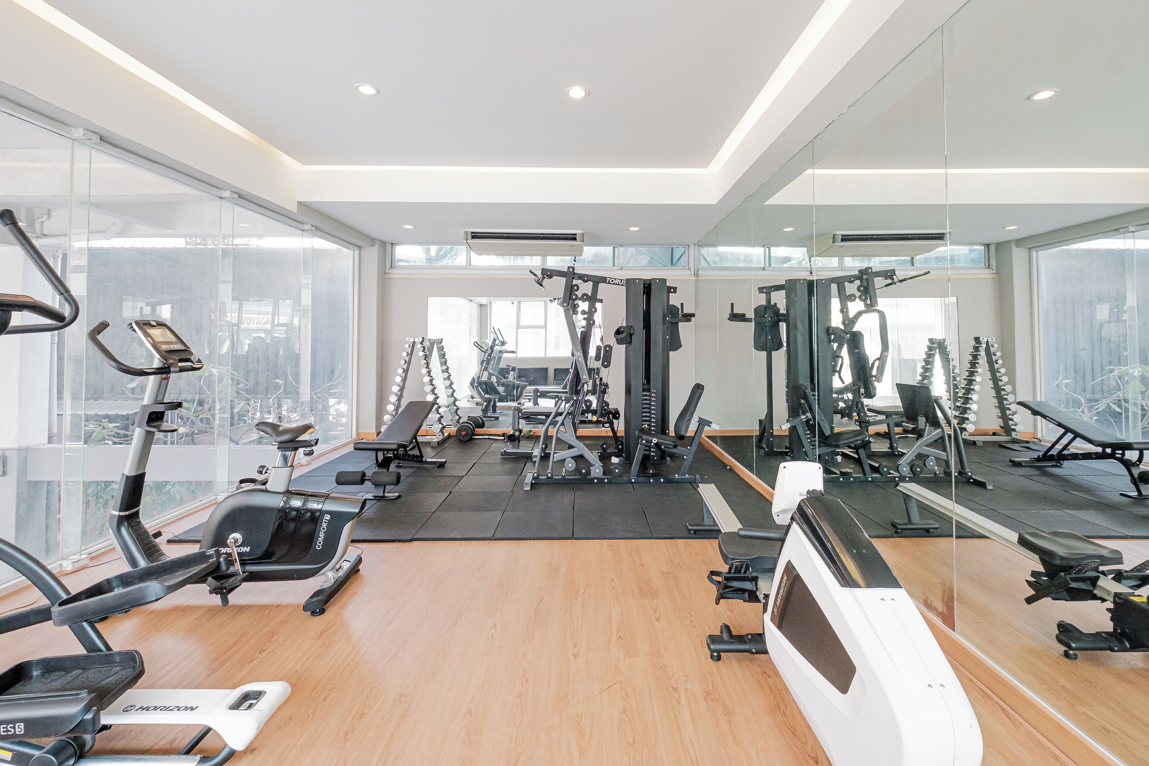 A modern gym with various exercise equipment, large windows, and a wooden floor.