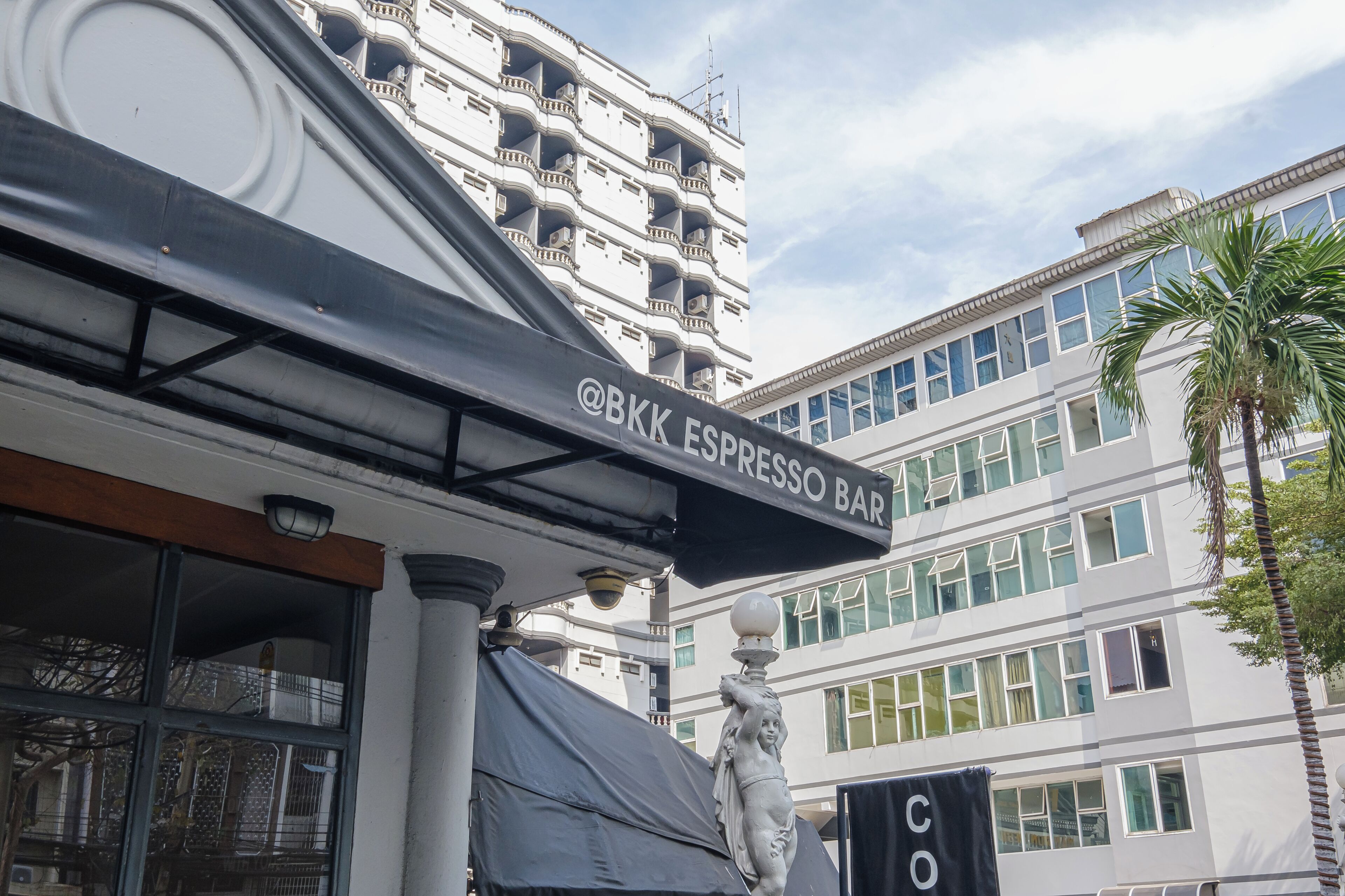 A sign for @BKK ESPRESSO BAR, a building with multiple balconies, and a palm tree.