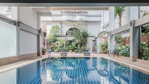 Indendørs swimmingpool med fliserede vægge og en central fontæne, omgivet af plantekasser og liggestole.