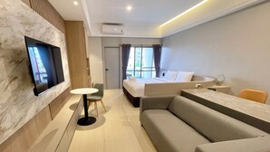 Et moderne hotelværelse med et fladskærms-tv monteret på væggen, en seng med hvide sengetøj, en grå sofa, et lille rundt bord og et stort vindue med gardiner.