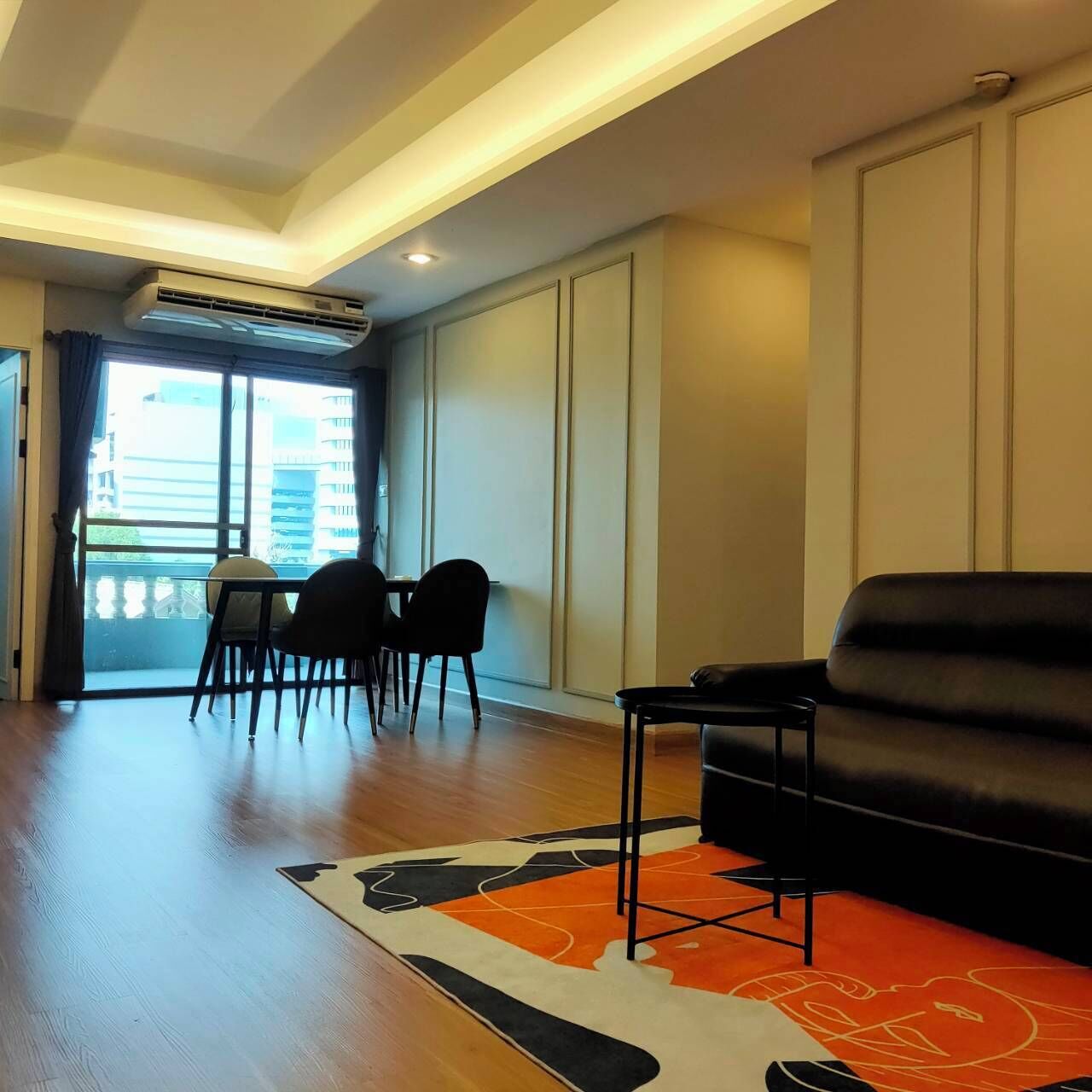 Uma sala de estar moderna com um sofá de couro preto, uma mesa de jantar com cadeiras e uma janela ampla com vista para edifícios.