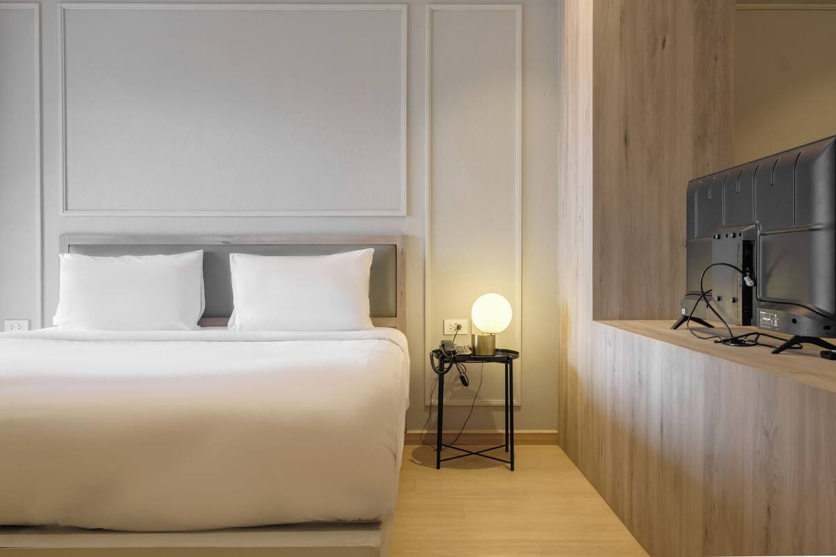 Una cama bien hecha con sábanas blancas, una mesita de noche con lámpara y un televisor de pantalla plana montado en un panel de madera.