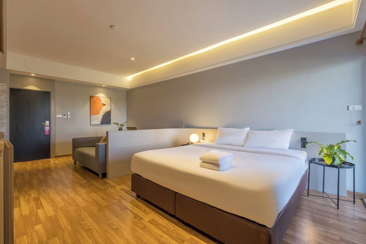 Una habitación de hotel moderna con una cama grande, una mesita de noche con lámpara, un pequeño rincón de estar y suelo de madera.