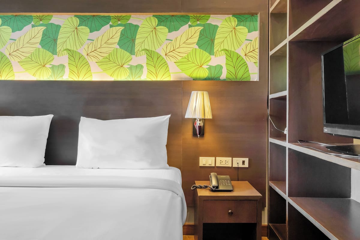 Habitación de hotel con cama, mesita de noche con lámpara y teléfono, televisor sobre una repisa y un papel tapiz con diseño de hojas.