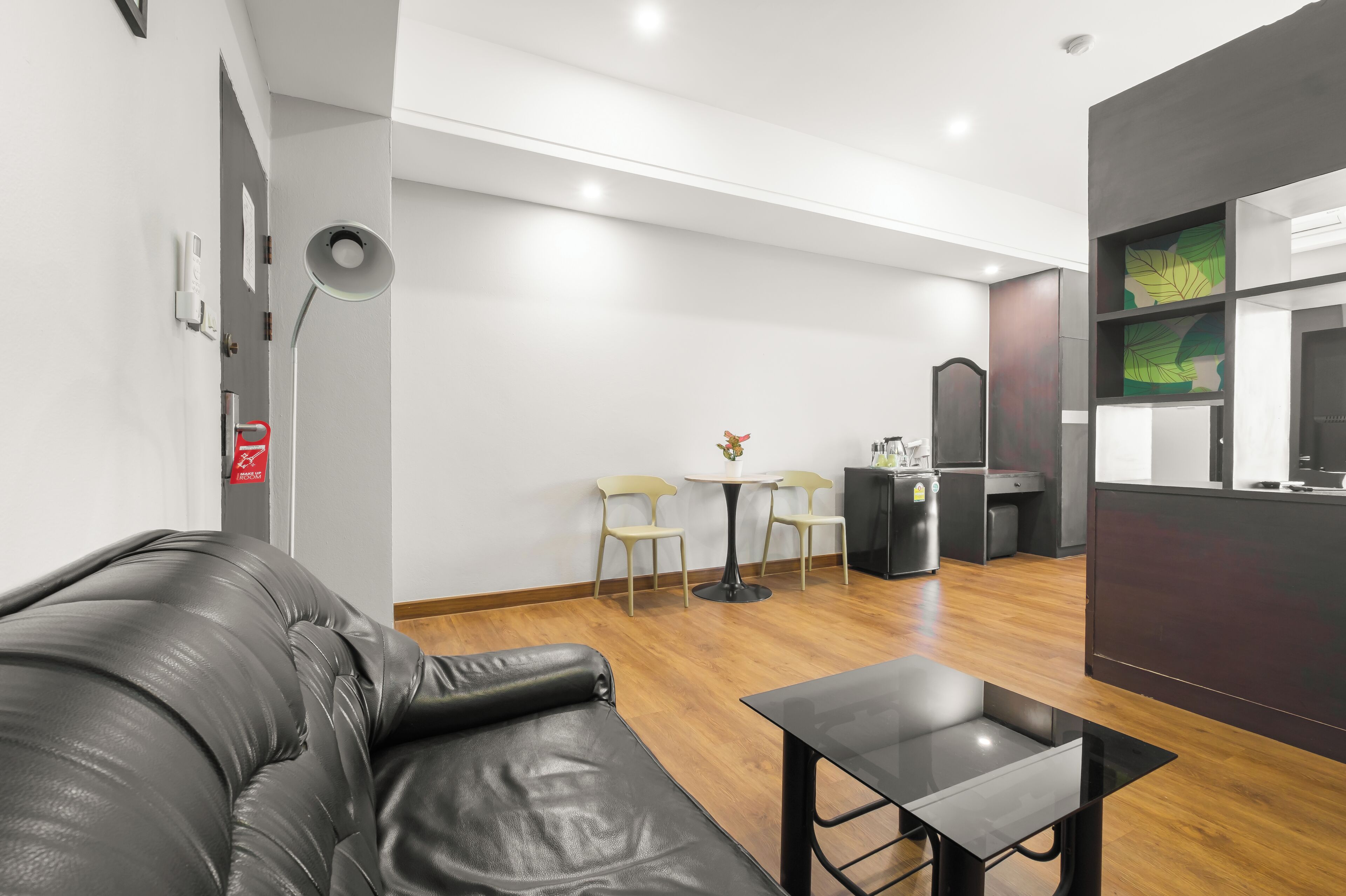 A modern hotel room with a leather sofa, a small table, two chairs, a round table with a vase, a minibar, and a large mirror.