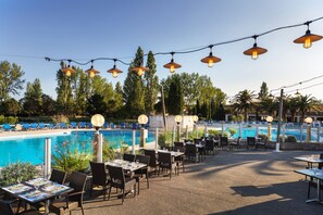 Zona de piscina exterior con sillones de descanso y mesas, rodeada de árboles y un cielo despejado.
