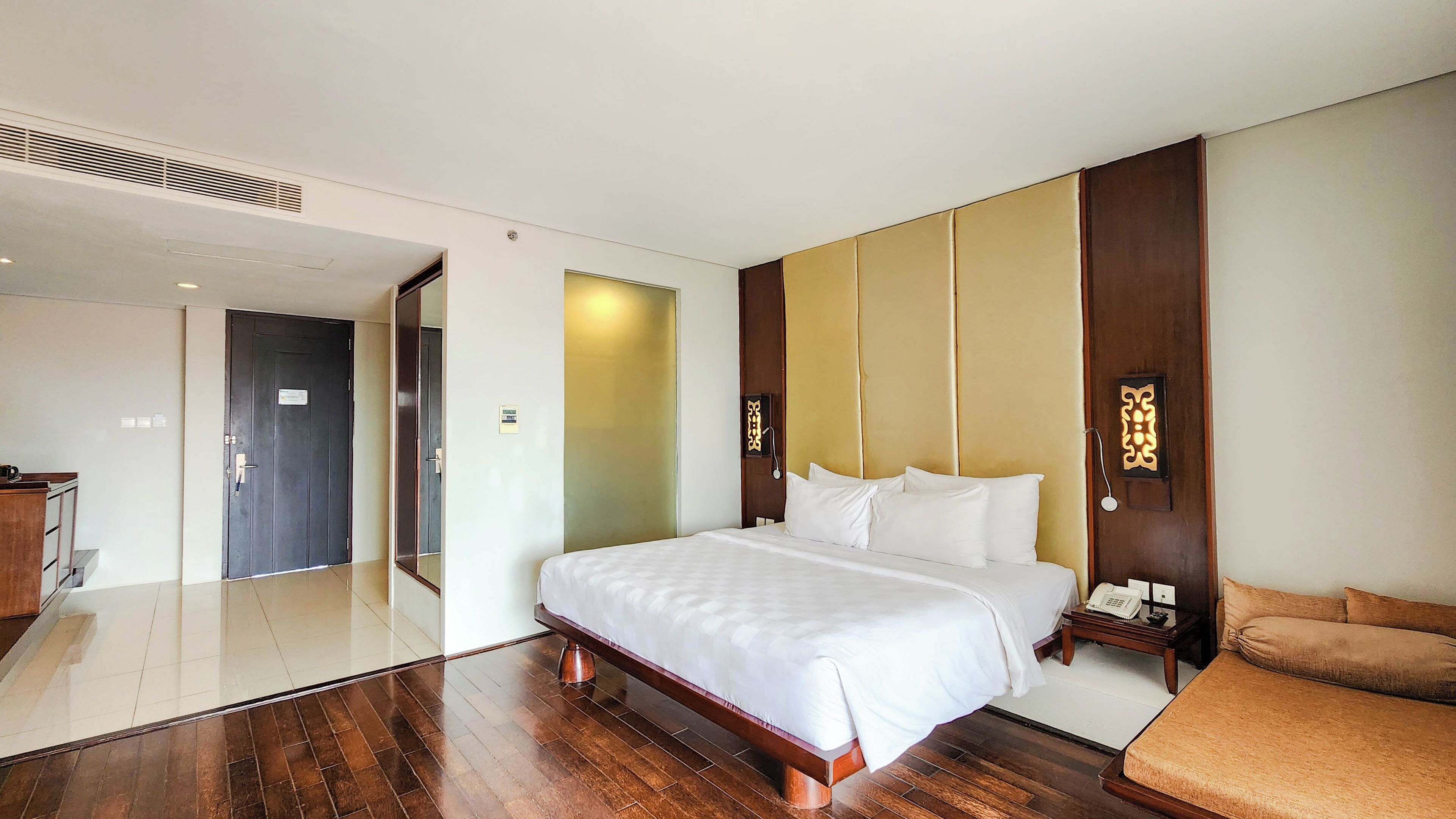 Una habitación de hotel con una cama grande, una mesita de noche de madera, una mesita con un teléfono y una pequeña zona de estar con un banco acolchado.