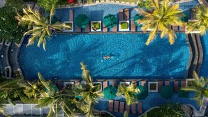 Une vue aérienne d’une grande piscine d’un bleu éclatant, entourée d’espaces de détente agrémentés de palmiers et de parasols.