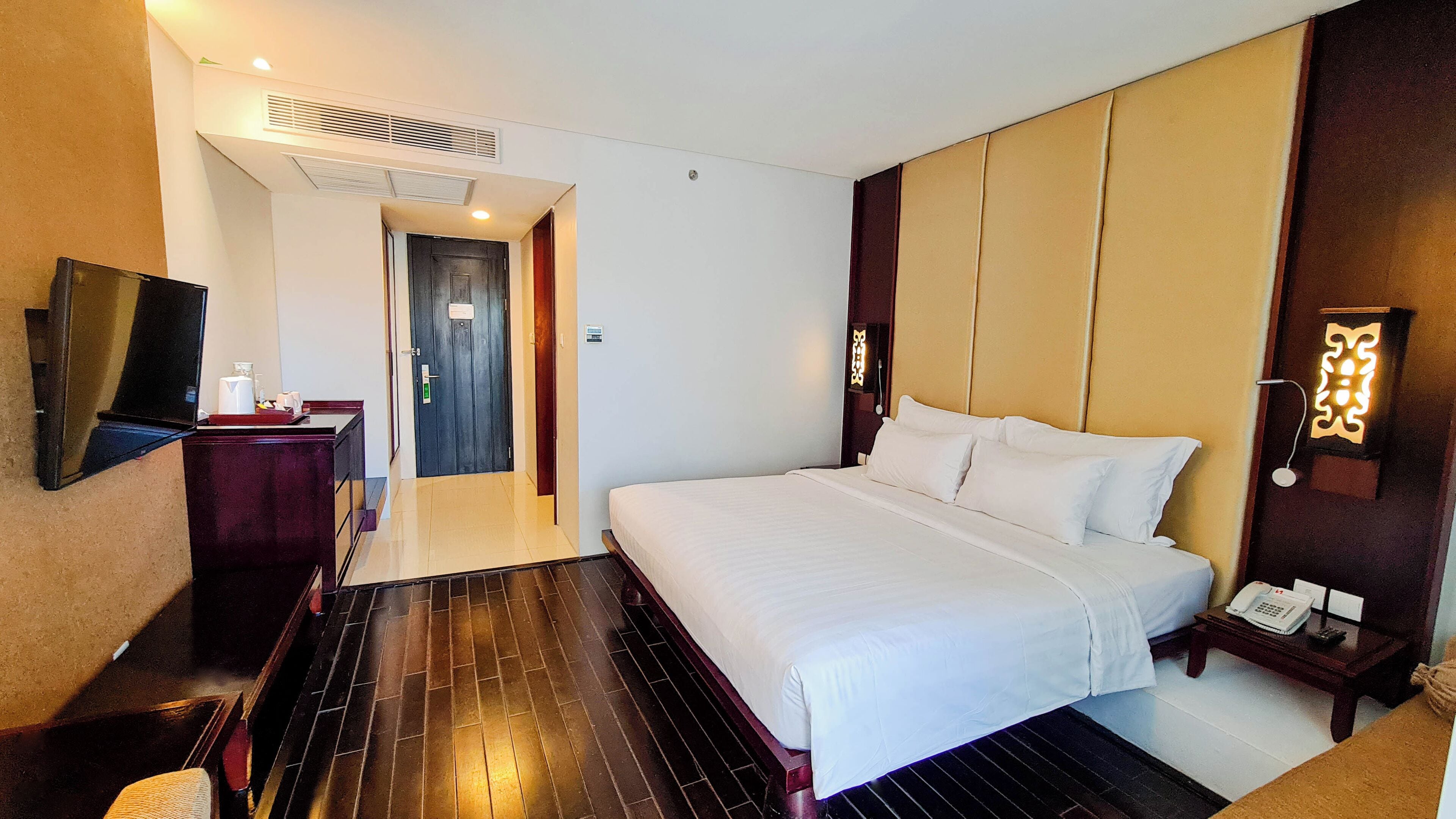 A hotel room with a large bed, a desk with a telephone, a TV, and a wall-mounted light fixture.