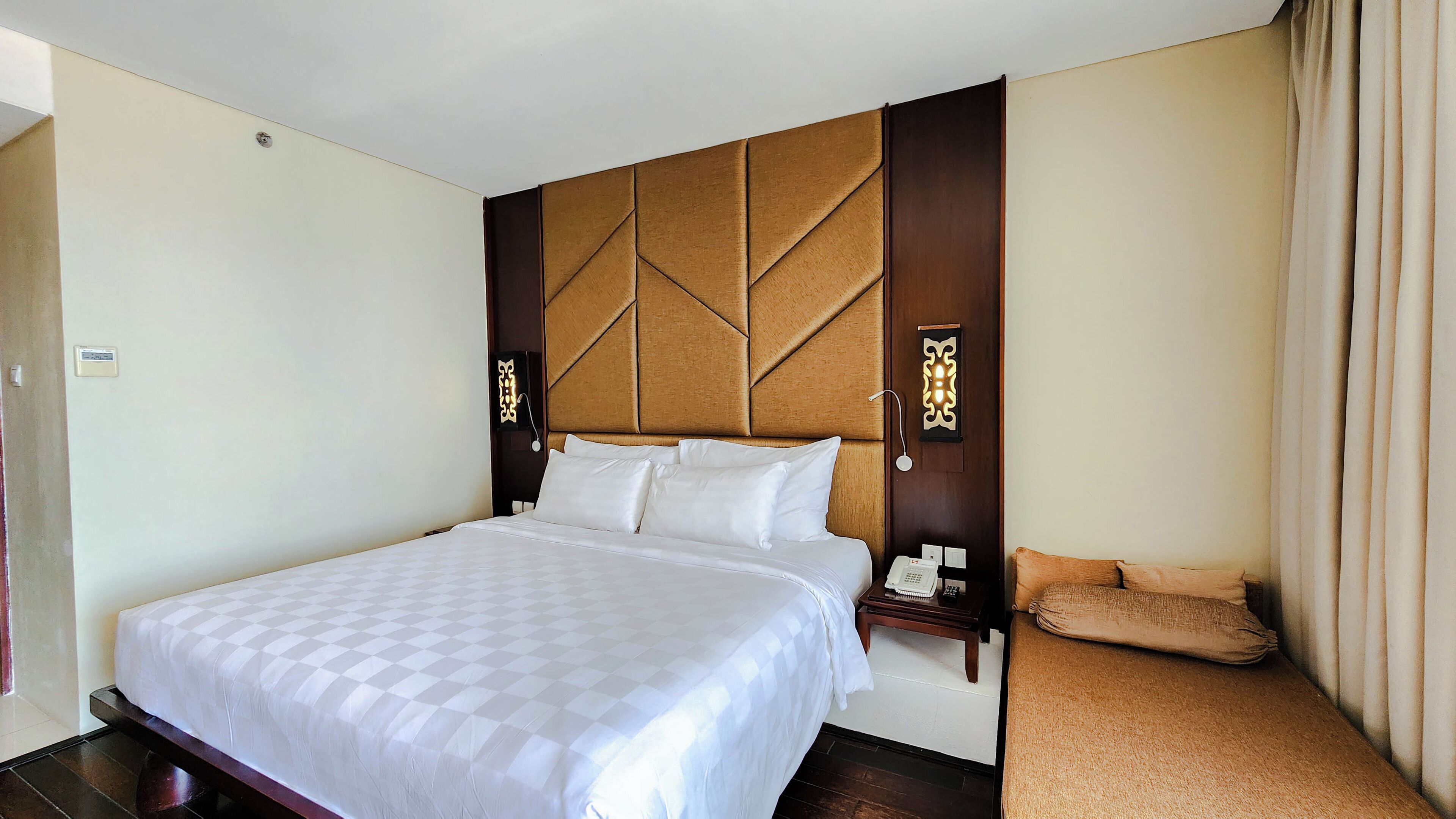 A hotel room with a large bed, a wooden headboard, a bedside table with a lamp, and a small sofa.