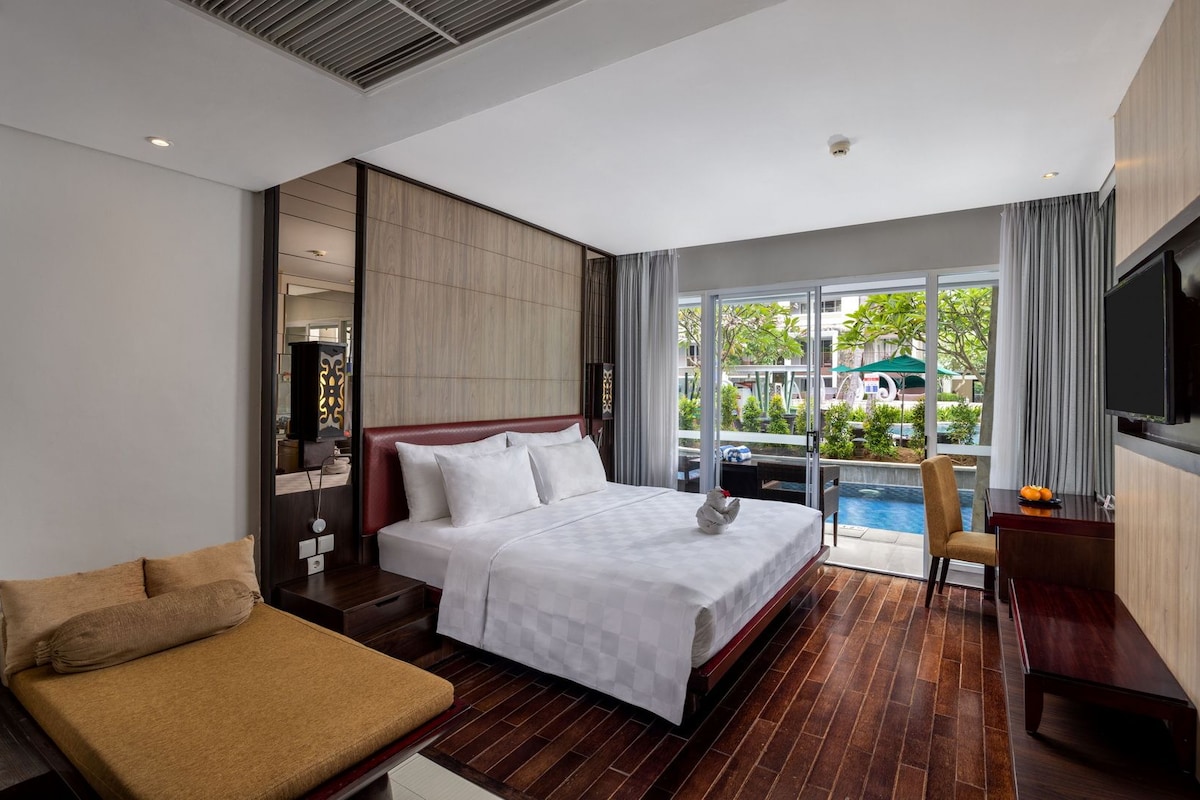 A hotel room with a bed, a sofa, a desk, a TV, and a view of an outdoor swimming pool.