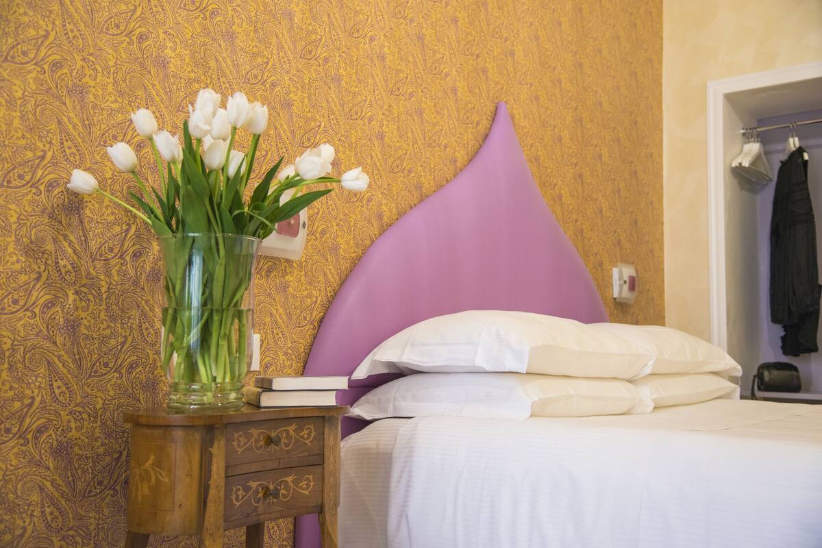 A bedroom with a bed, a nightstand, a vase with white flowers, and a patterned wallpaper.