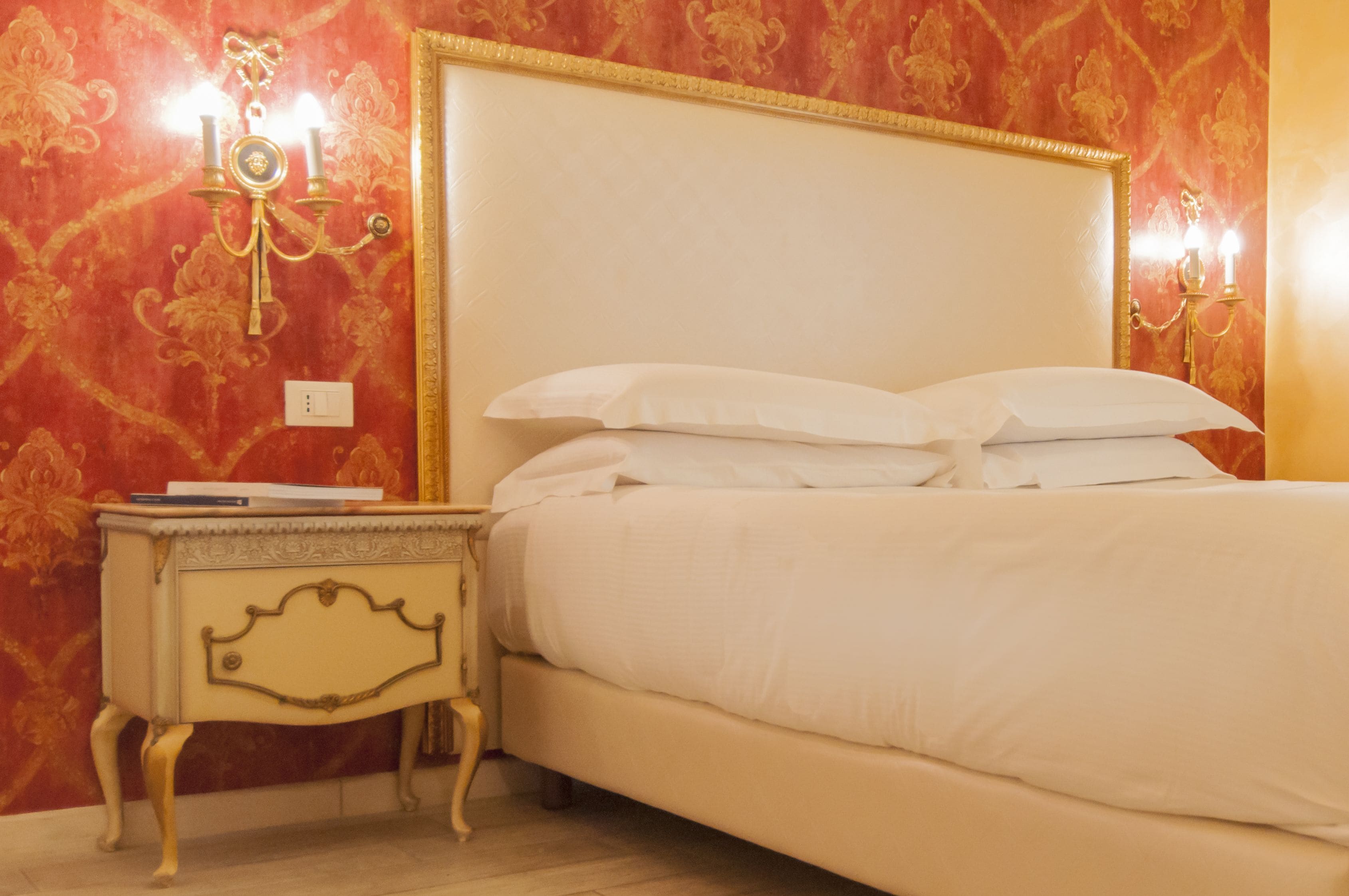 A bed with white linens in a room with red patterned wallpaper and gold trim.