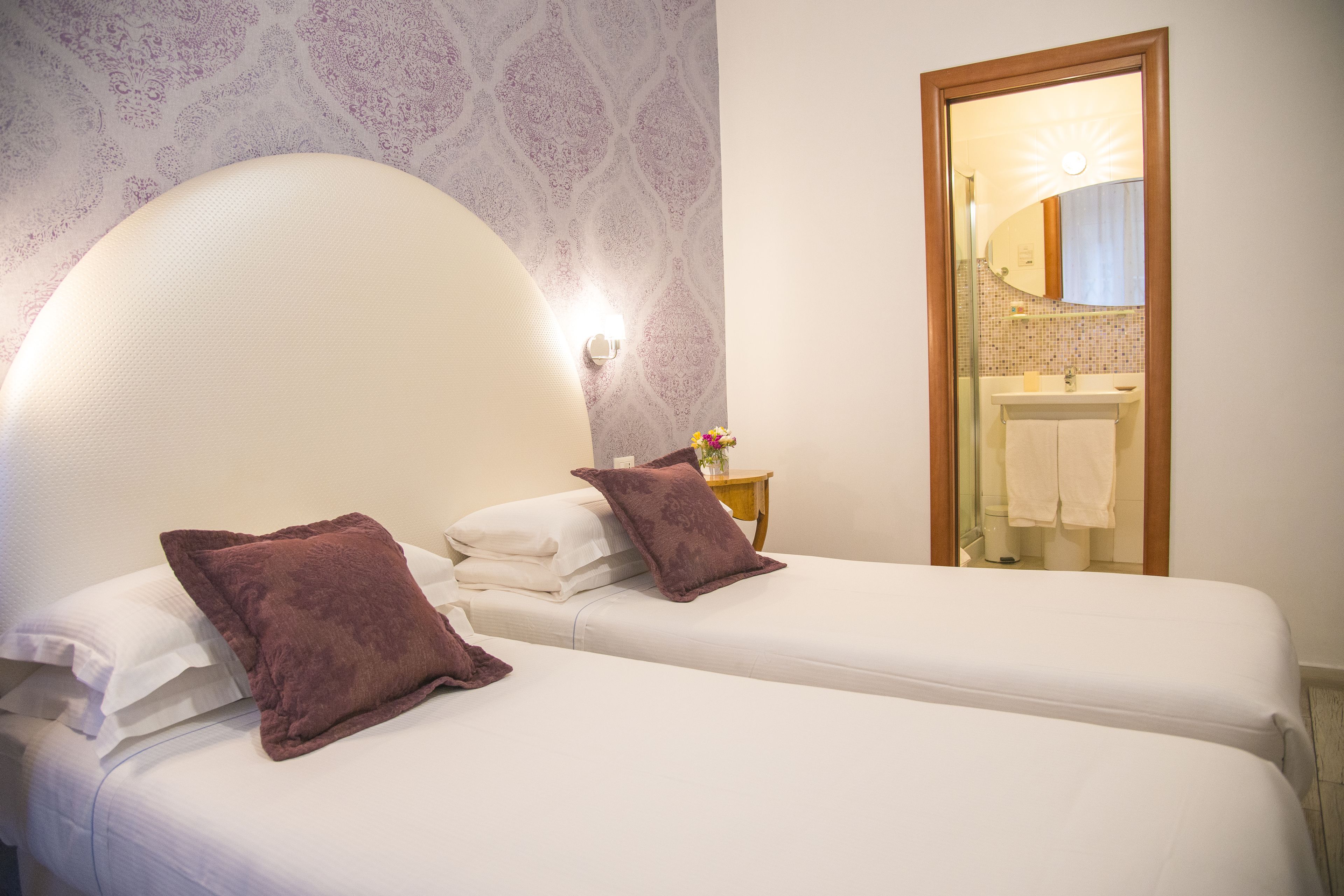A hotel room with two beds, a large mirror, a nightstand with a lamp, and a bathroom visible through an open door.