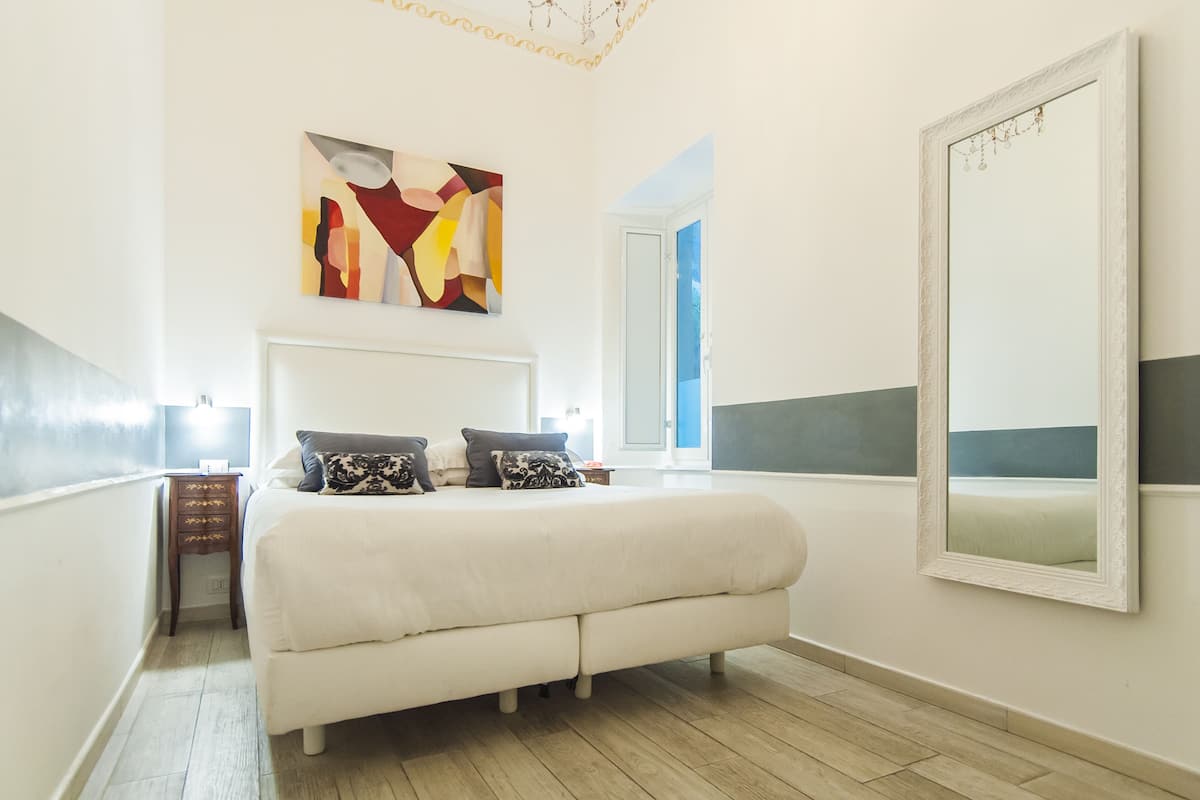 A bedroom with a large bed, a nightstand, a wall-mounted mirror, and a painting.
