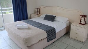 A neatly made bed with a striped blanket and pillows, a bedside table with a lantern, and a view of the outdoors through a window with curtains. - Surfers Del Rey (Surfers Paradise)