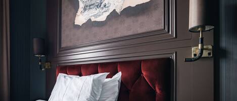 Una cama bien hecha con ropa blanca y un cabecero tapizado en rojo. Sobre la cama, un cuadro enmarcado que representa un naufragio.