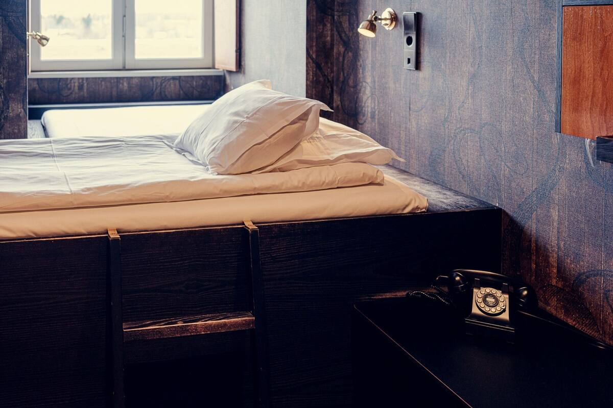 A neatly made bed with white linens in a room with a wooden wall and a window.