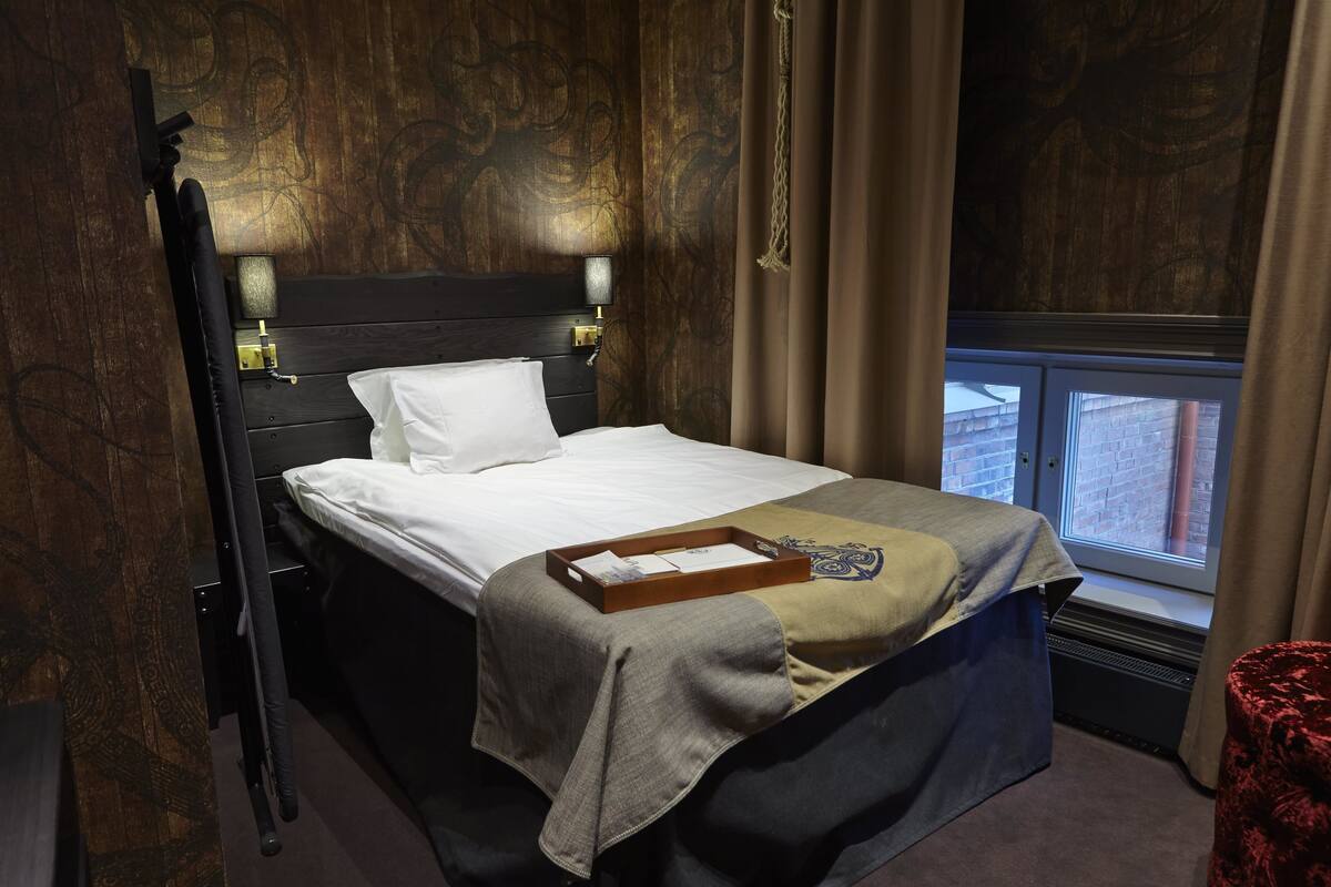 A neatly made bed with a tray and a remote control, a bedside table with a lamp, and a window with curtains.