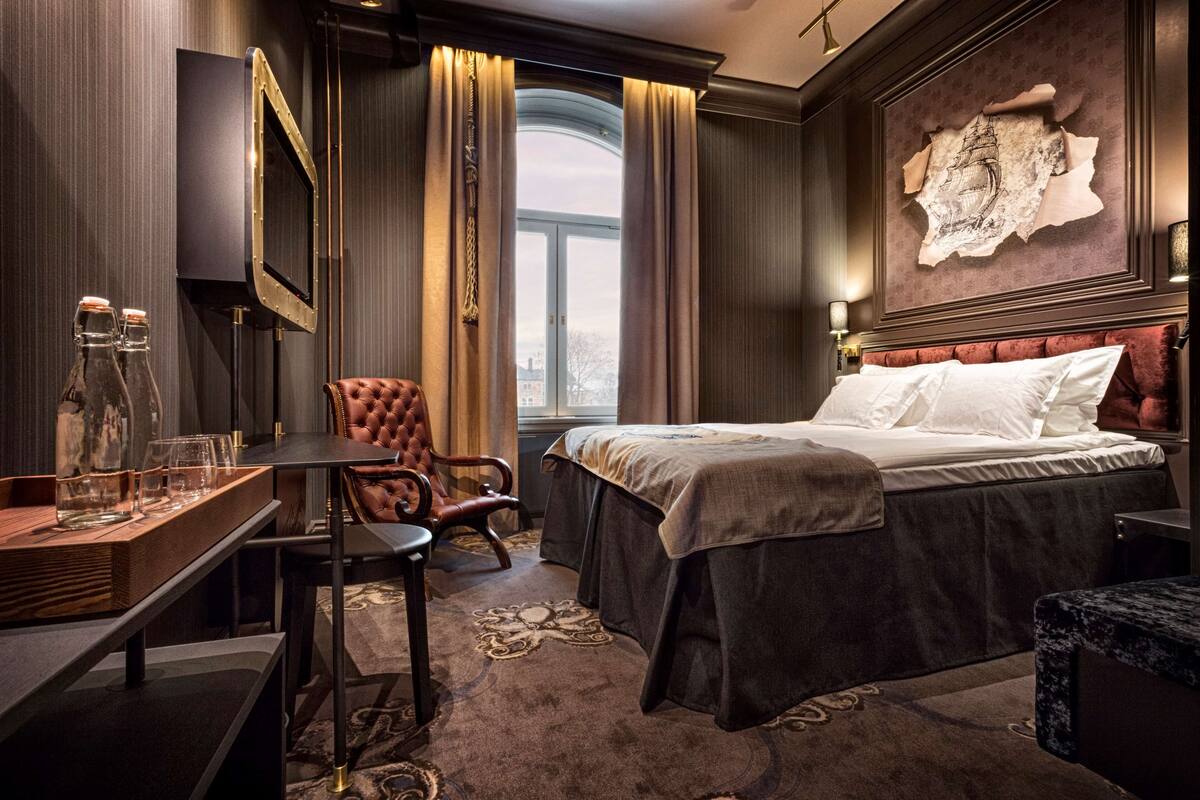 A hotel room with a large bed, a desk with a chair, a window with curtains, and a wall-mounted TV.