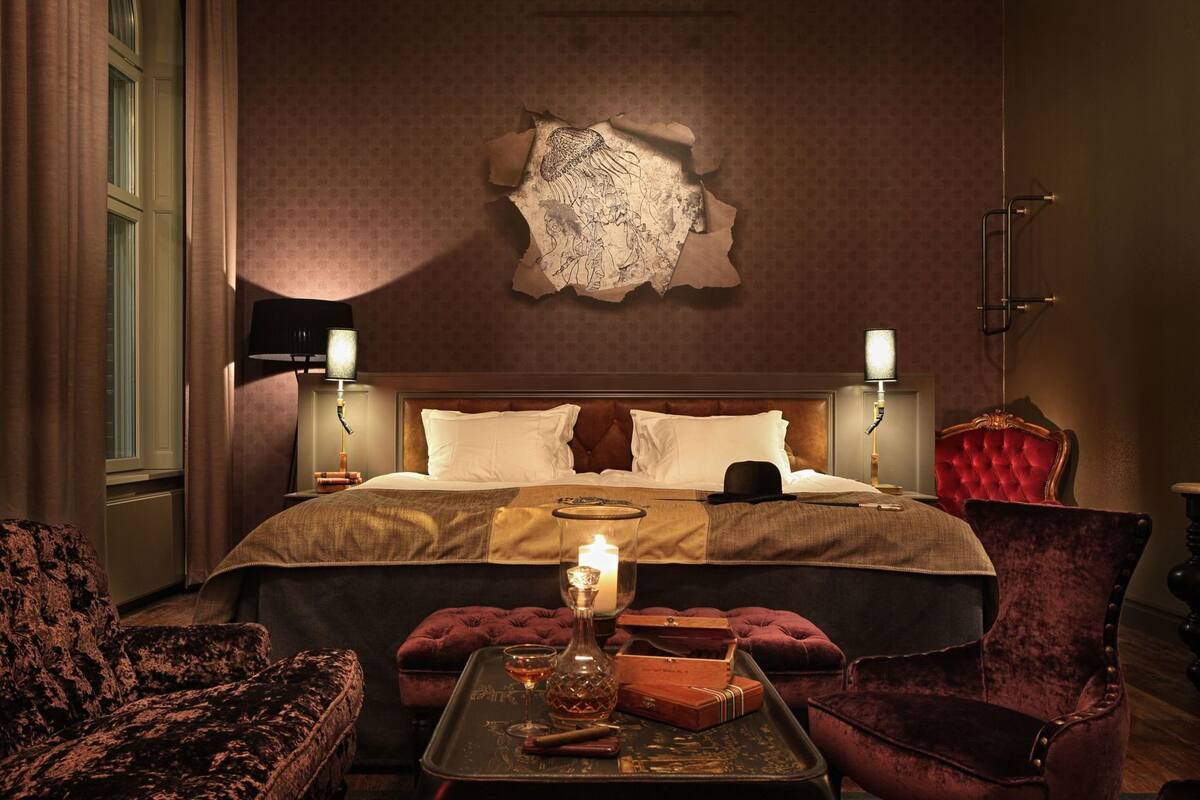A hotel room with a large bed, two bedside lamps, a red velvet armchair, a small table with a candle, and a decorative wall piece.