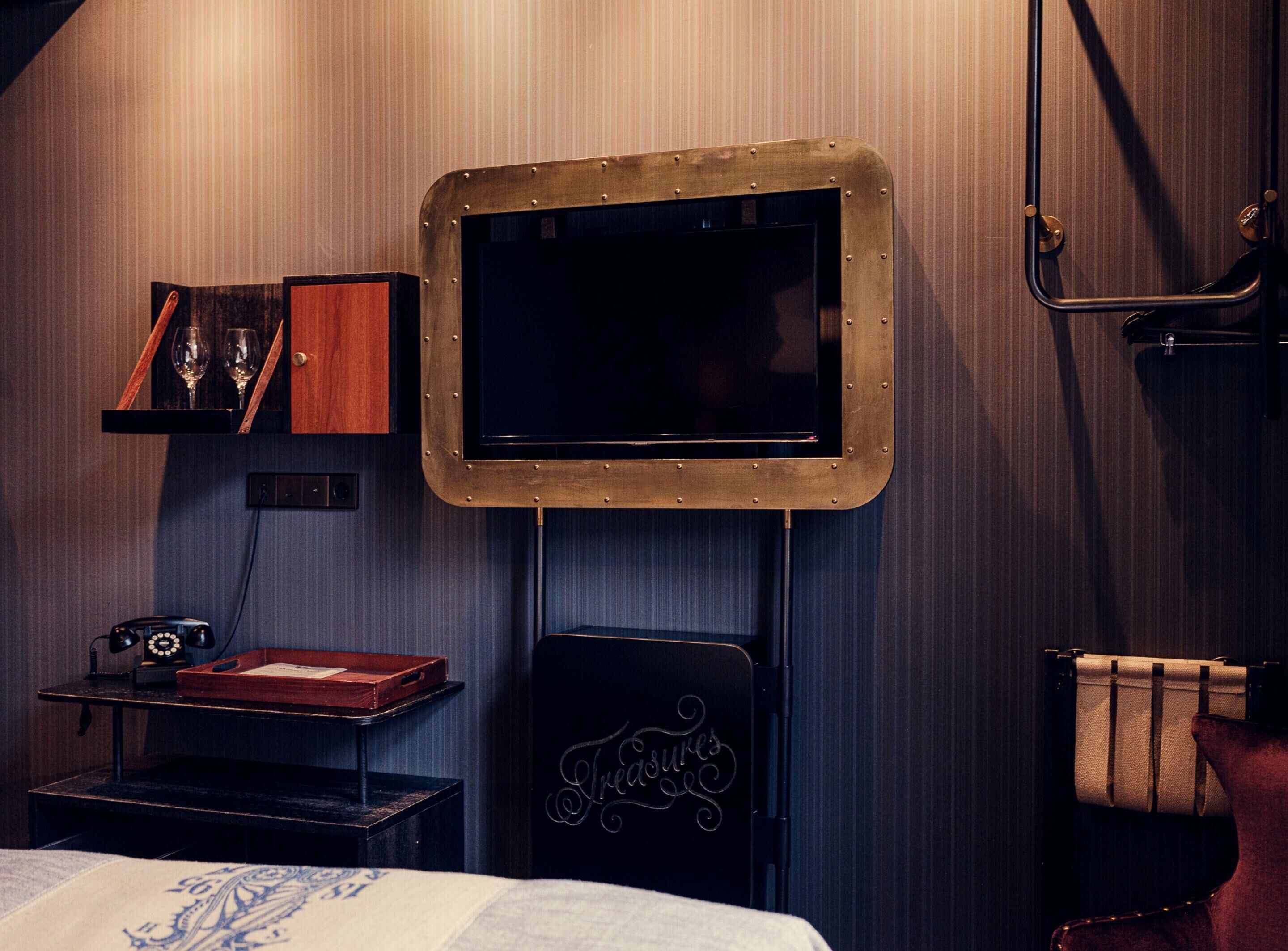 A hotel room with a mounted television, a vintage telephone, a small table with a red tray, and a decorative item with the word "Treasures".