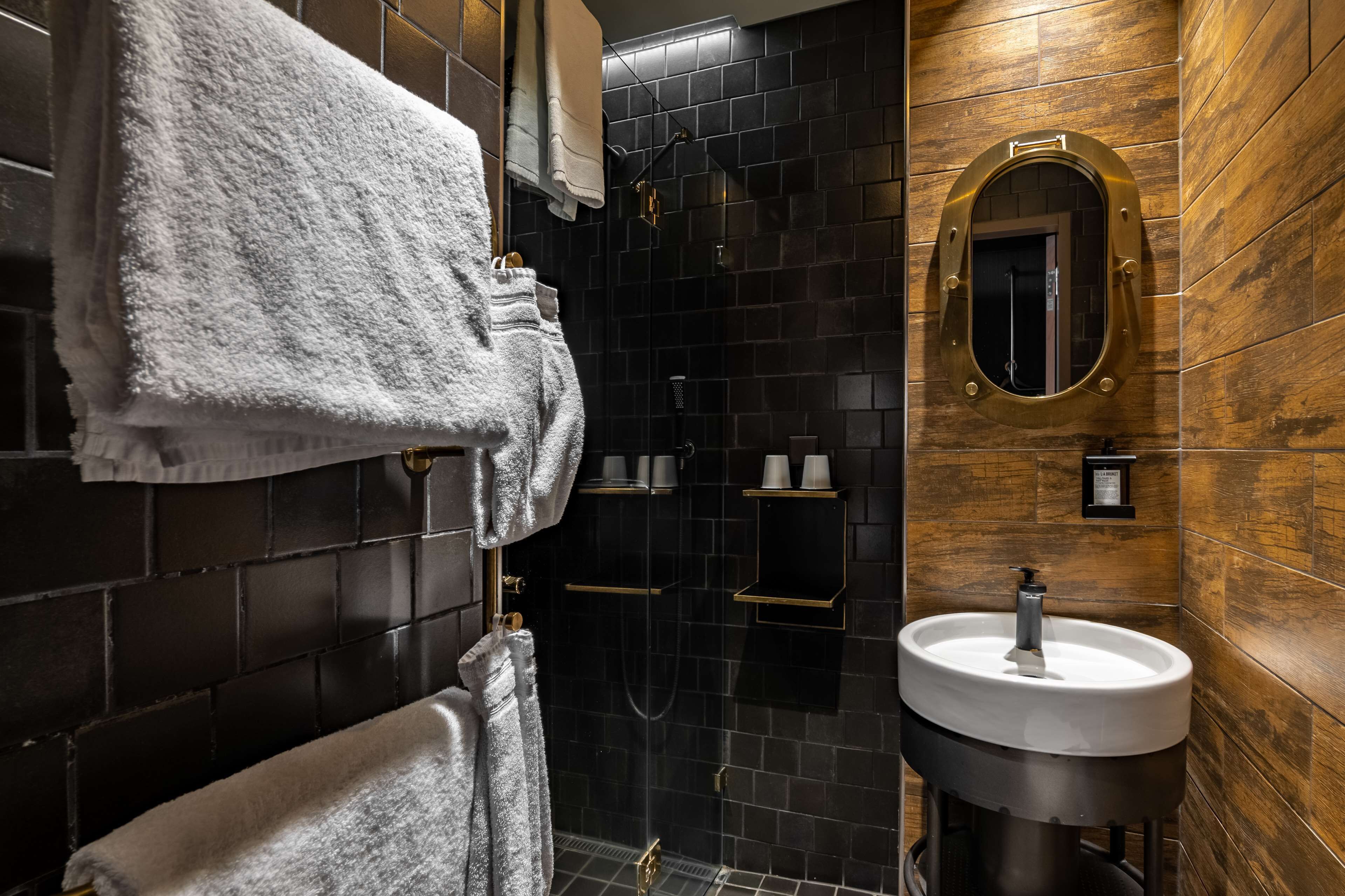 A modern bathroom with a black tiled shower, a white sink with a gold faucet, and wooden wall panels.