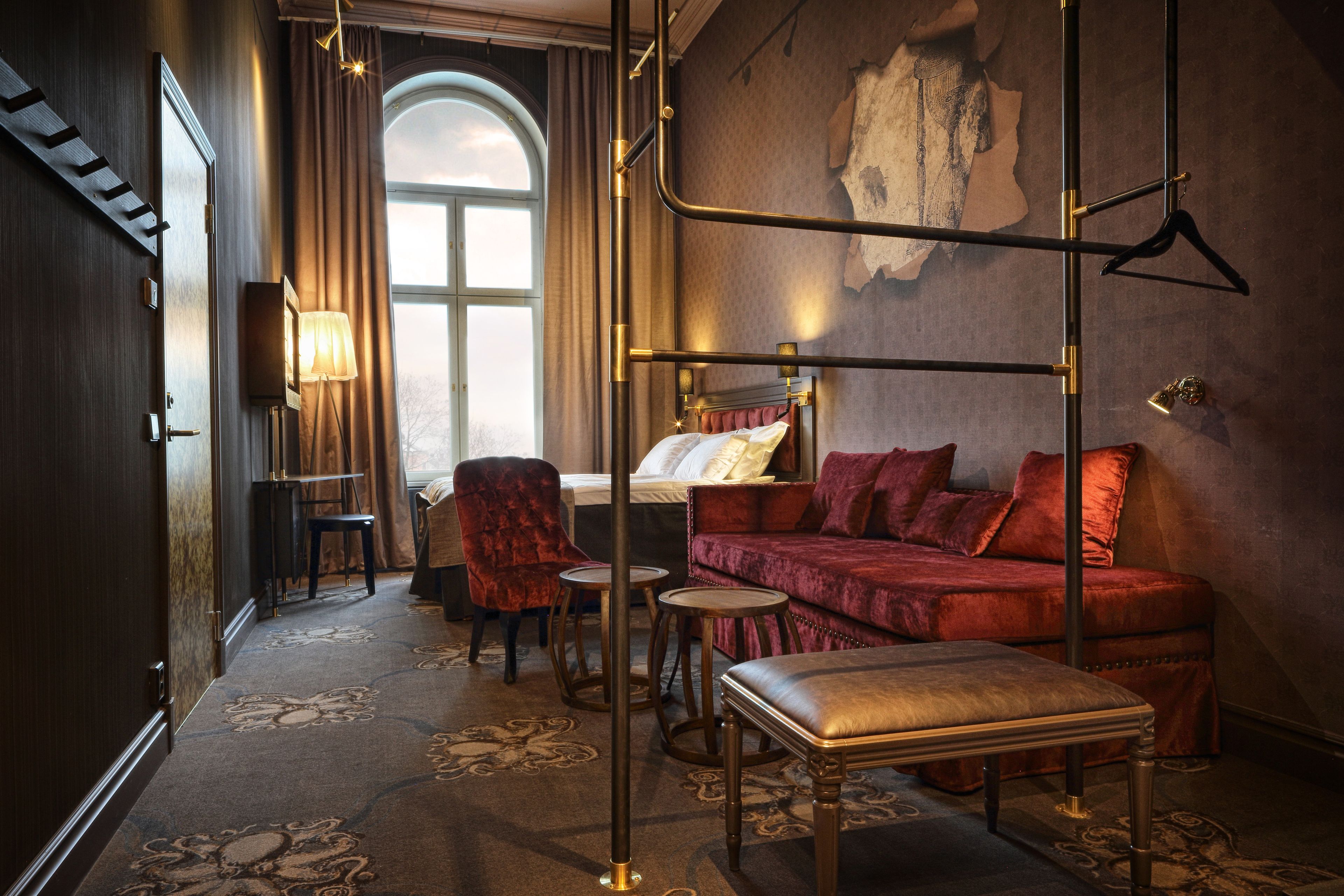Una habitación de hotel con una cama, un sofá de terciopelo rojo, una mesita, una silla y una lámpara colgante.
