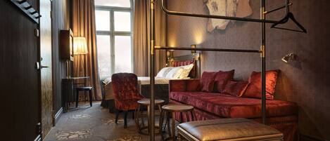 Una habitación de hotel con una cama, un sofá de terciopelo rojo, una mesita, una silla y una lámpara colgante.