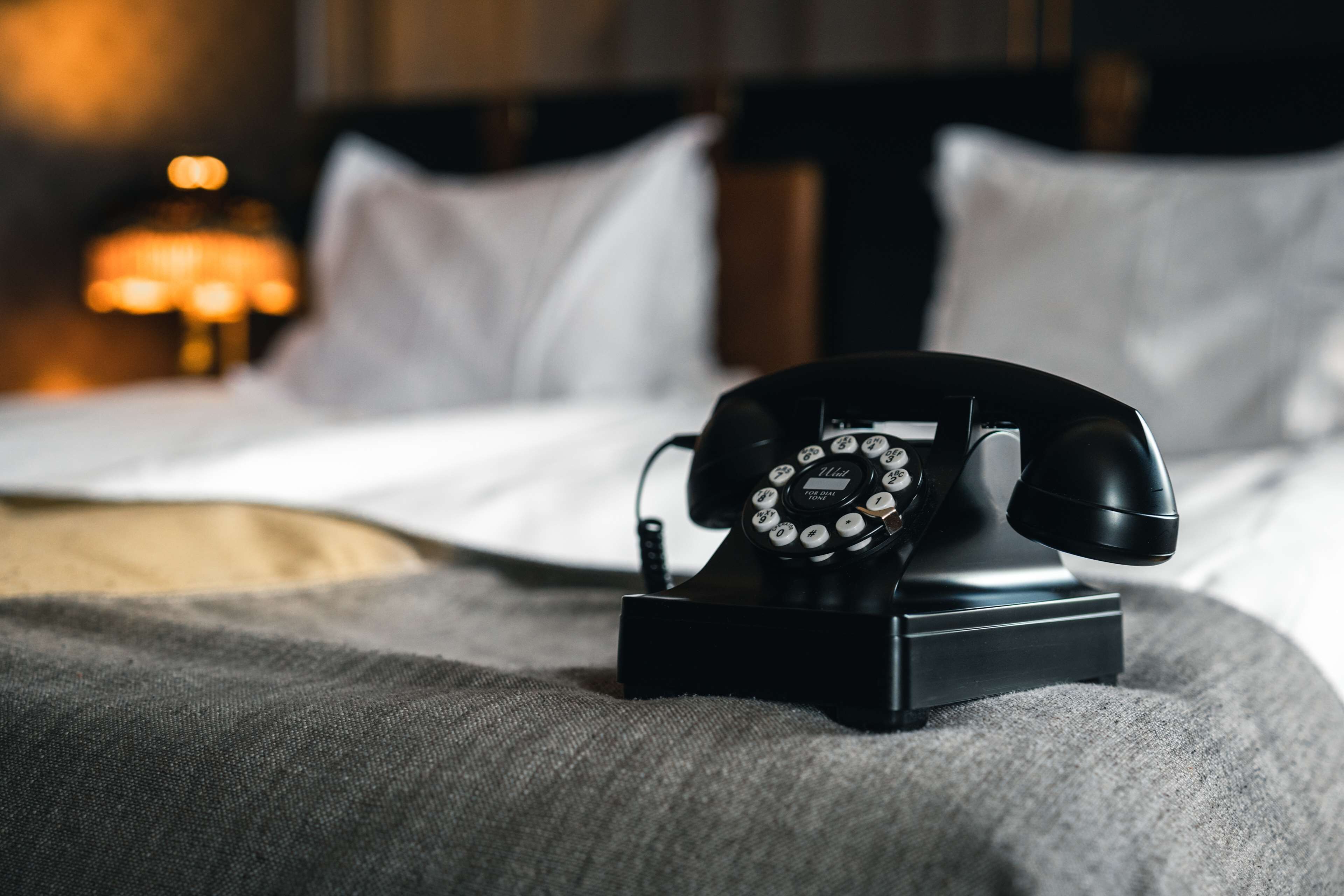A black rotary telephone on a bedside table in a hotel room with a bed and a lamp in the background.