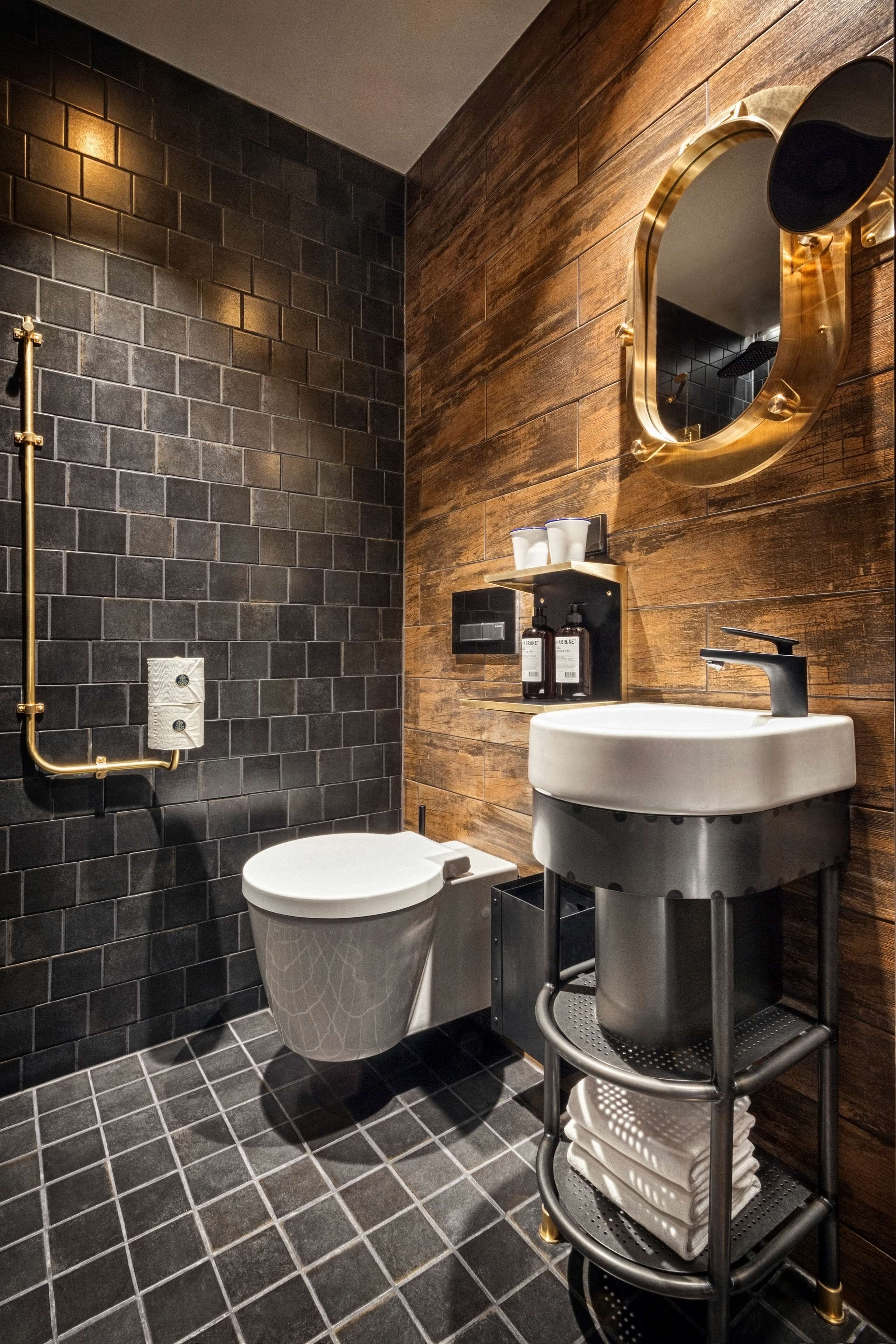 A modern bathroom with a black tiled wall, a wooden accent wall, a white toilet, a sink with a gold faucet, and a mirror with a gold frame.