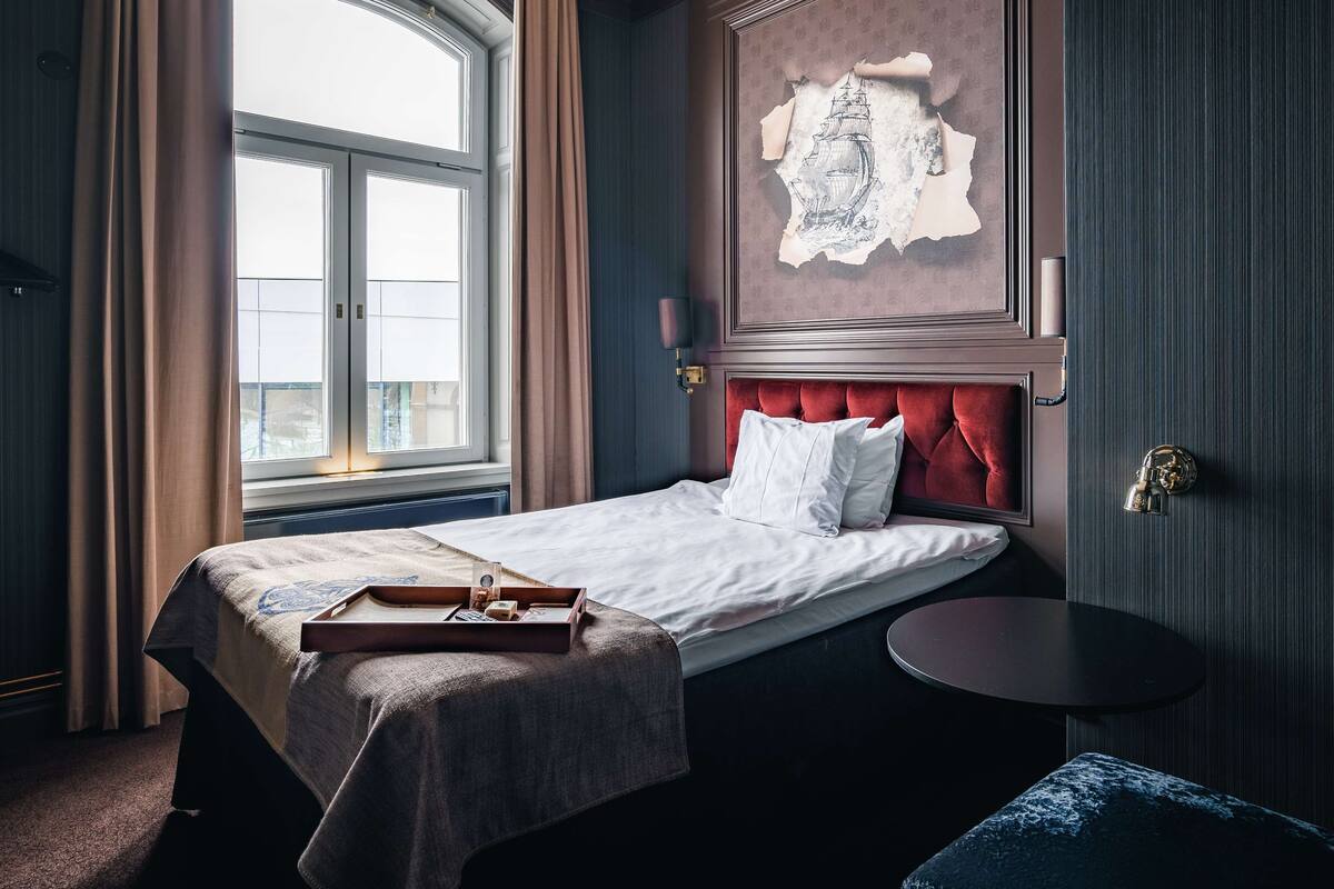 A hotel room with a large window, a bed with a red headboard, a small round table, and a wall-mounted lamp.