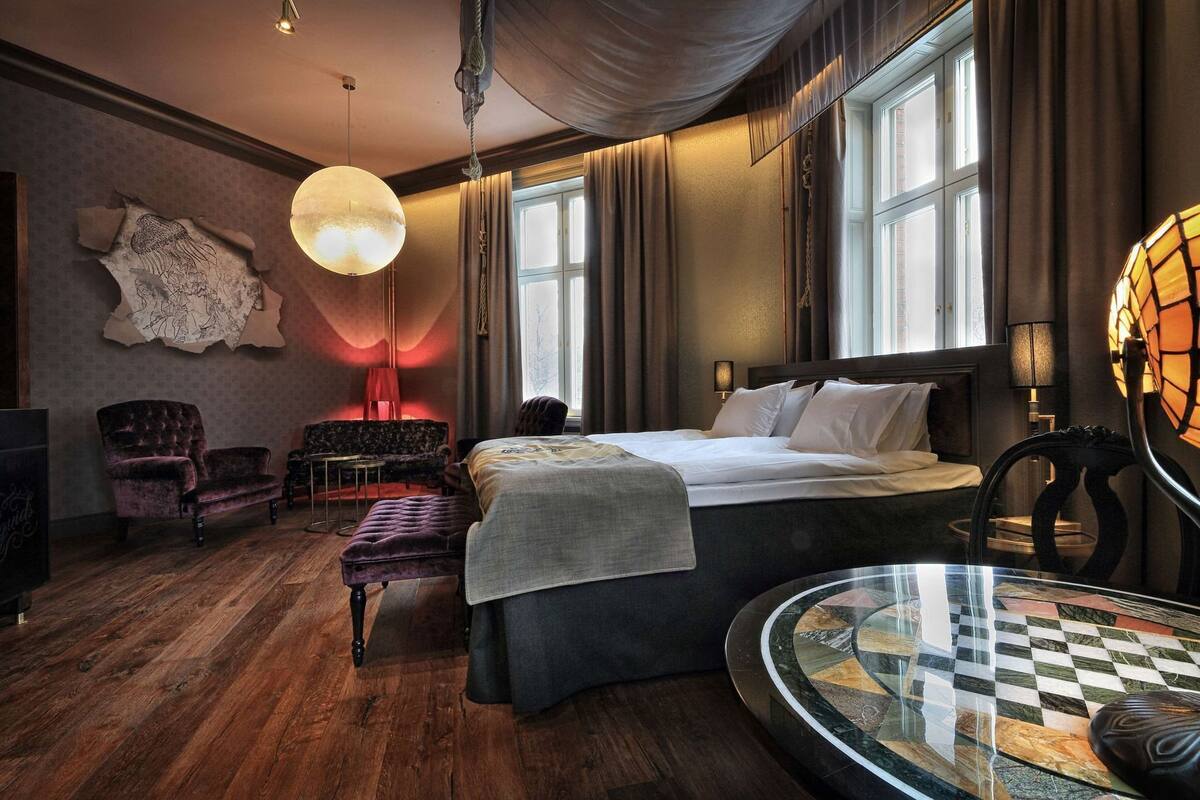 A hotel room with a large bed, a round glass table, a sofa, and a chandelier.