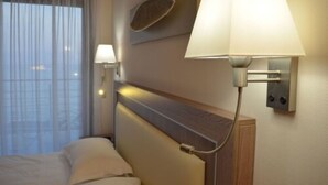 A hotel room with a bed, a nightstand, a wall-mounted lamp, and a window with sheer curtains. - Hotel Villa Azur (Villeneuve-Loubet)